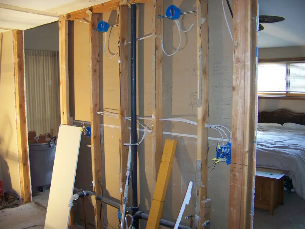 Before — Master bathroom remodel. Before: gutted to studs with exposed wiring/plumbing visible from adjoining bedroom. After: finished double-sink granite vanity with maple cabinets and tall linen tower in same alcove.
