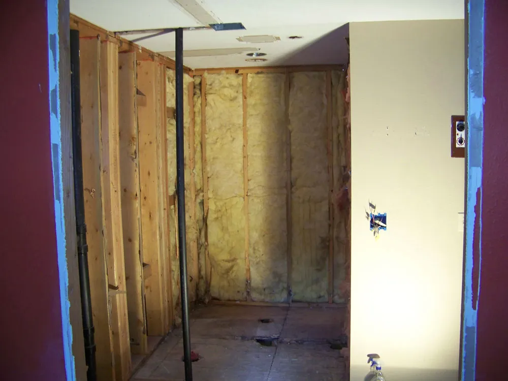 Before — Master bathroom shower area. Before: framed shower stall with pink/yellow insulation visible through opening from red bedroom wall. After: finished walk-in tile shower with stone-look tile, dual showerheads, bench, and corner linen cabinet.