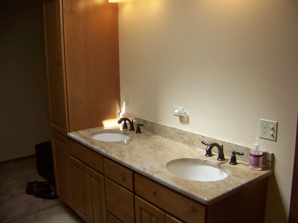 After — Master bathroom remodel. Before: gutted to studs with exposed wiring/plumbing visible from adjoining bedroom. After: finished double-sink granite vanity with maple cabinets and tall linen tower in same alcove.