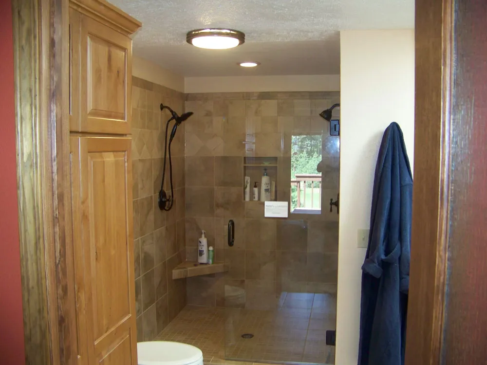 After — Master bathroom shower area. Before: framed shower stall with pink/yellow insulation visible through opening from red bedroom wall. After: finished walk-in tile shower with stone-look tile, dual showerheads, bench, and corner linen cabinet.
