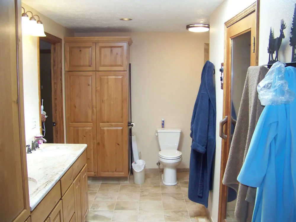 After — Master bathroom toilet/floor area. Before: subfloor torn up, pink/yellow insulation in walls, water damage visible. After: full bathroom with toilet, knotty alder cabinets, tile floor, recessed lighting, sauna door.