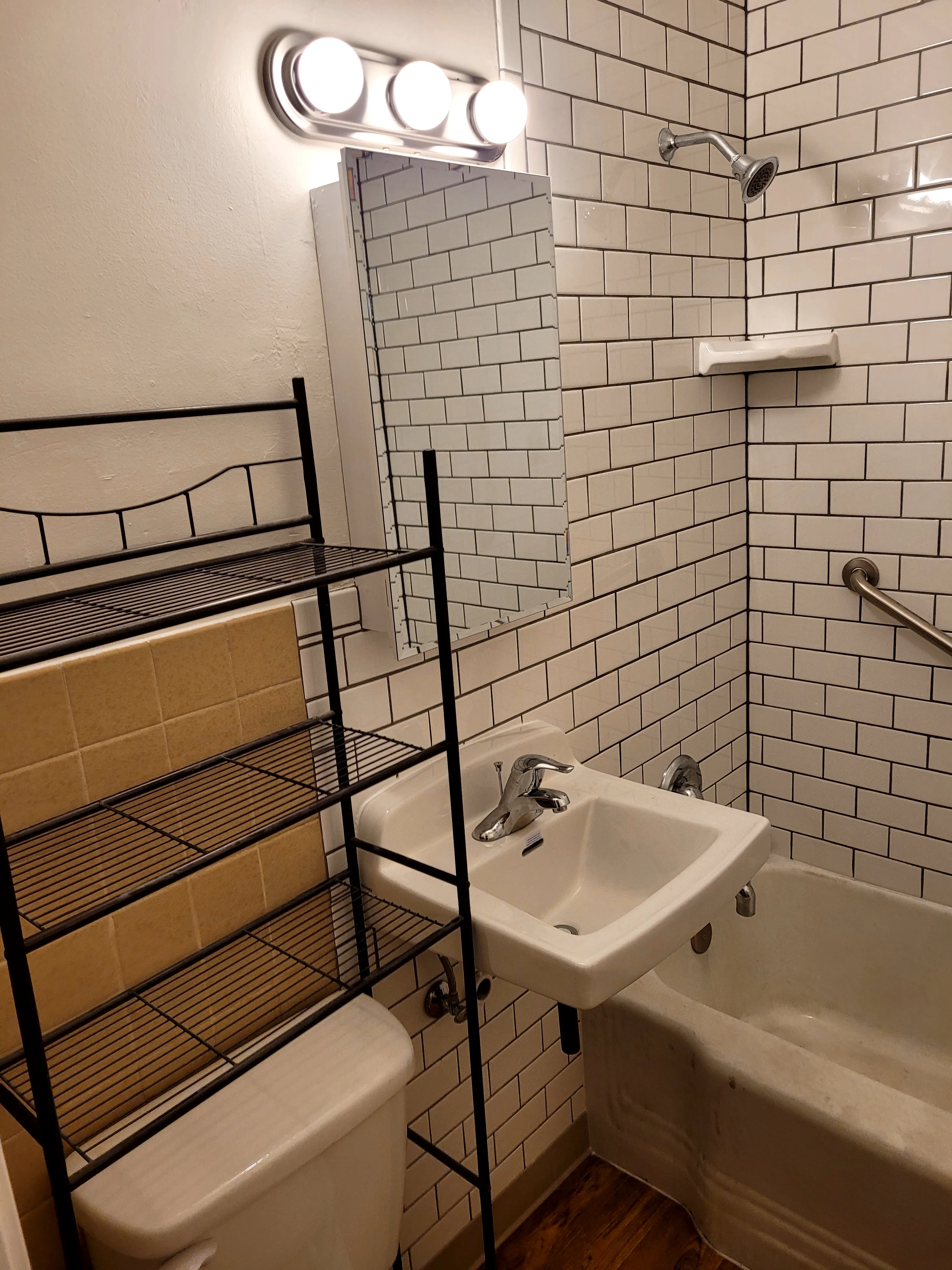 After — Same small bathroom — toilet/floor view. Before: scuffed brown vinyl tile floor with stained tub apron. After: white subway tile walls, new white toilet, hardwood floor.