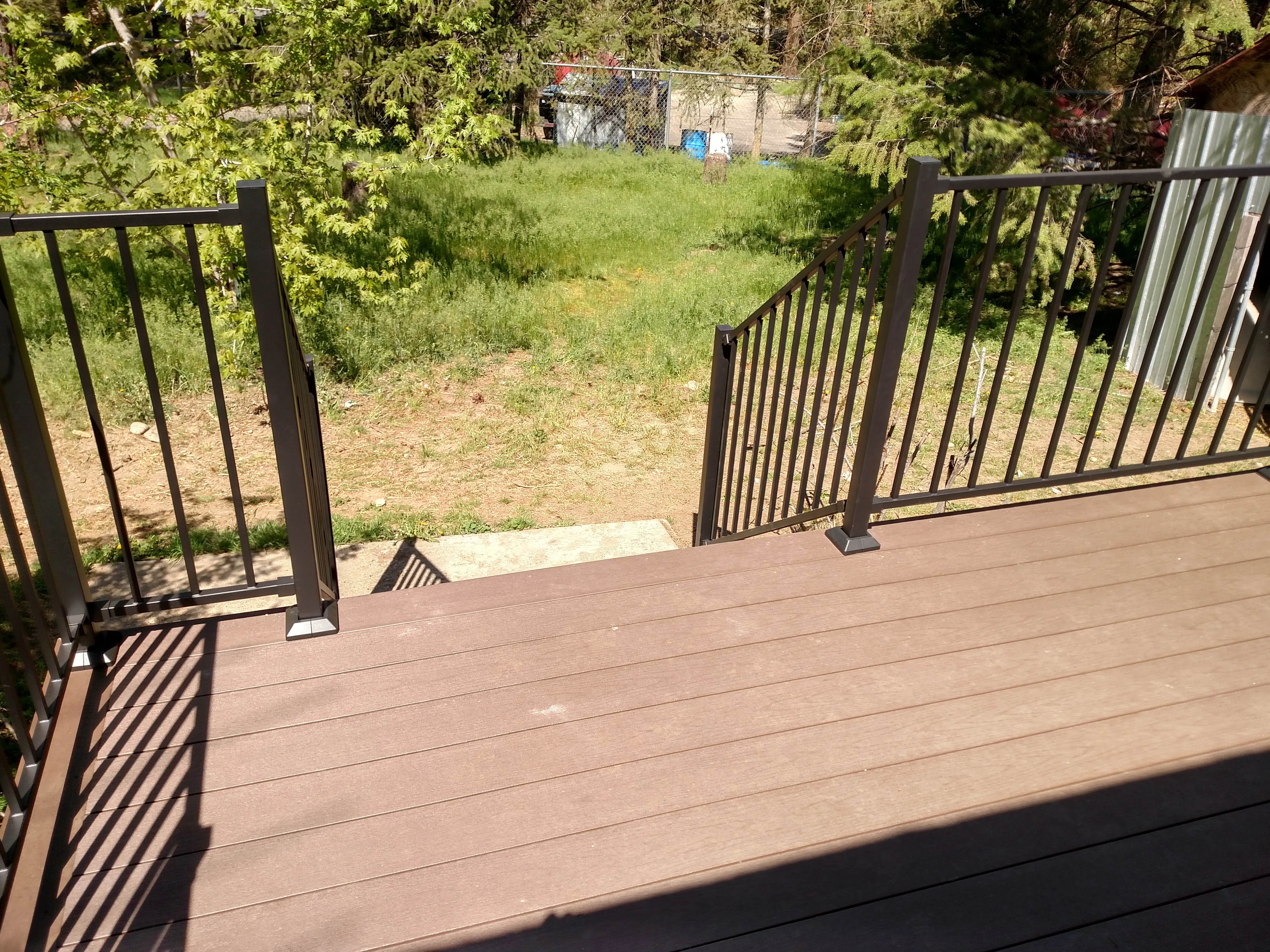 After — Backyard deck rebuild. Before: rotting wood deck with broken lattice skirt and weathered steps. After: new composite deck boards with black metal balusters and stairs leading to backyard.