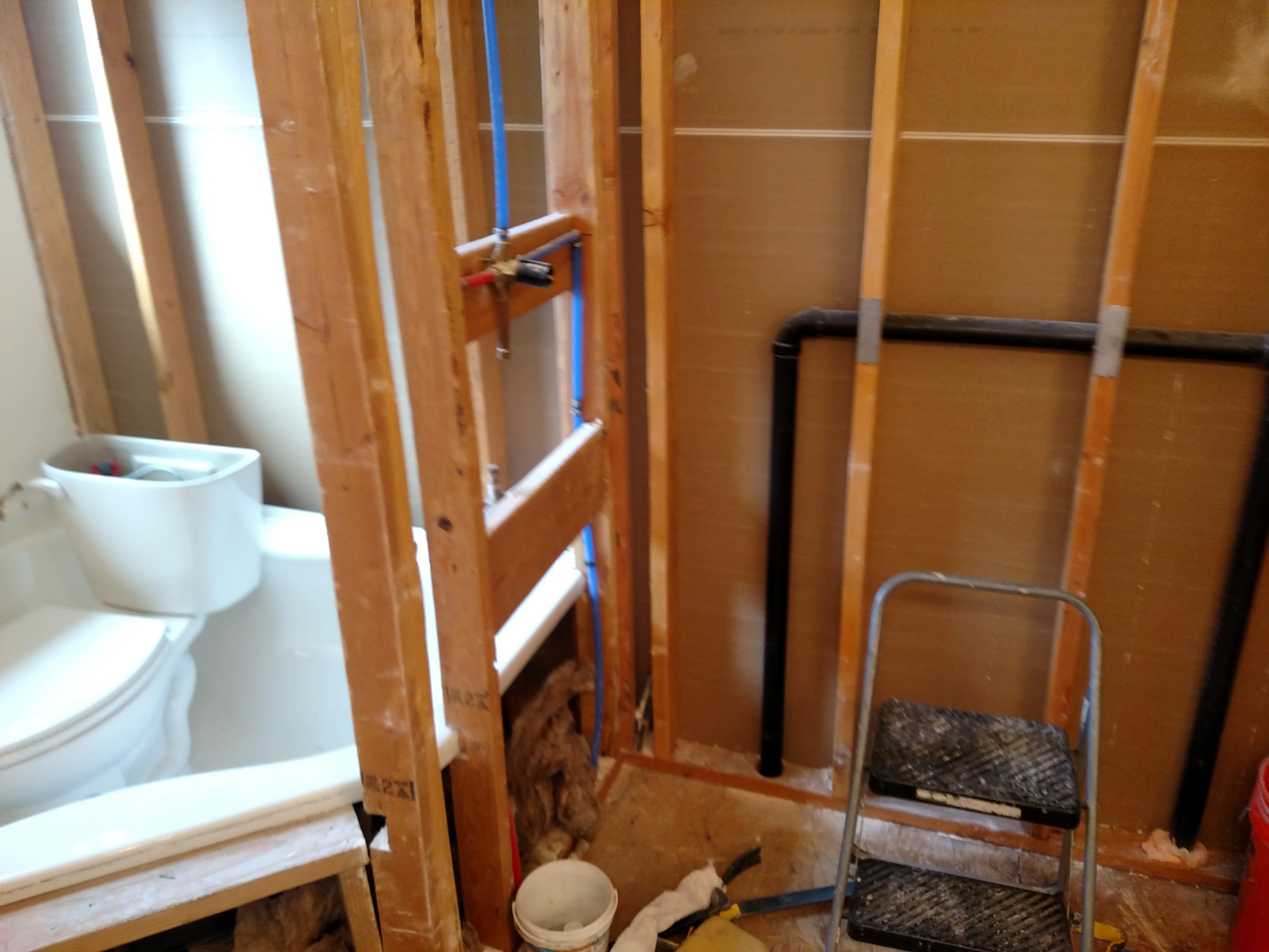 Before — Forest-view bathroom remodel. Before: gutted to studs with new blue PEX plumbing, old corner tub left in place. During: cement board (Durock) shower base built up under window. After: finished walk-in shower with white linear tile, pebble-stone shower floor, blue vanity, granite counter, knotty alder door, and the same forest window.