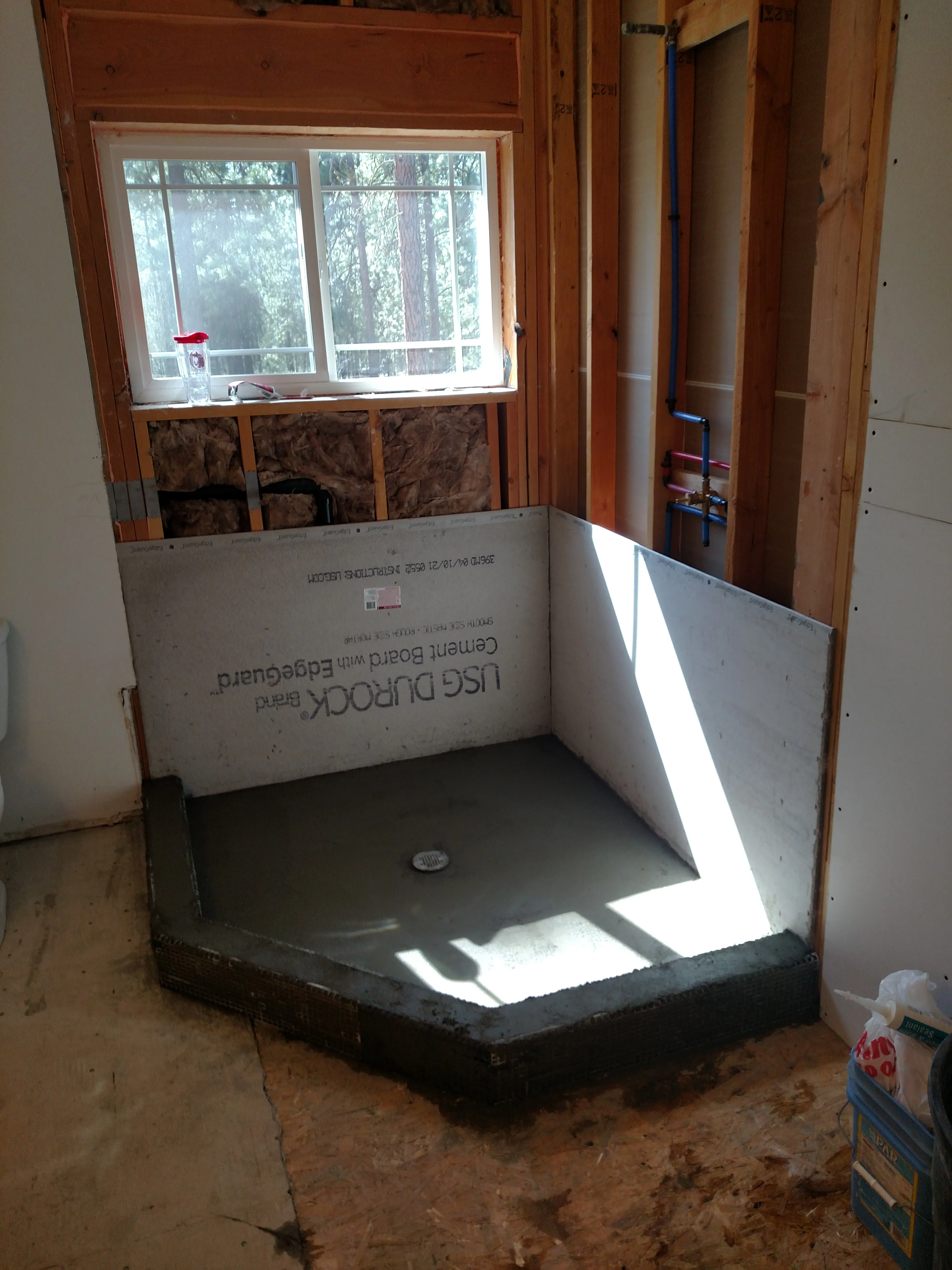 In Progress — Forest-view bathroom remodel. Before: gutted to studs with new blue PEX plumbing, old corner tub left in place. During: cement board (Durock) shower base built up under window. After: finished walk-in shower with white linear tile, pebble-stone shower floor, blue vanity, granite counter, knotty alder door, and the same forest window.