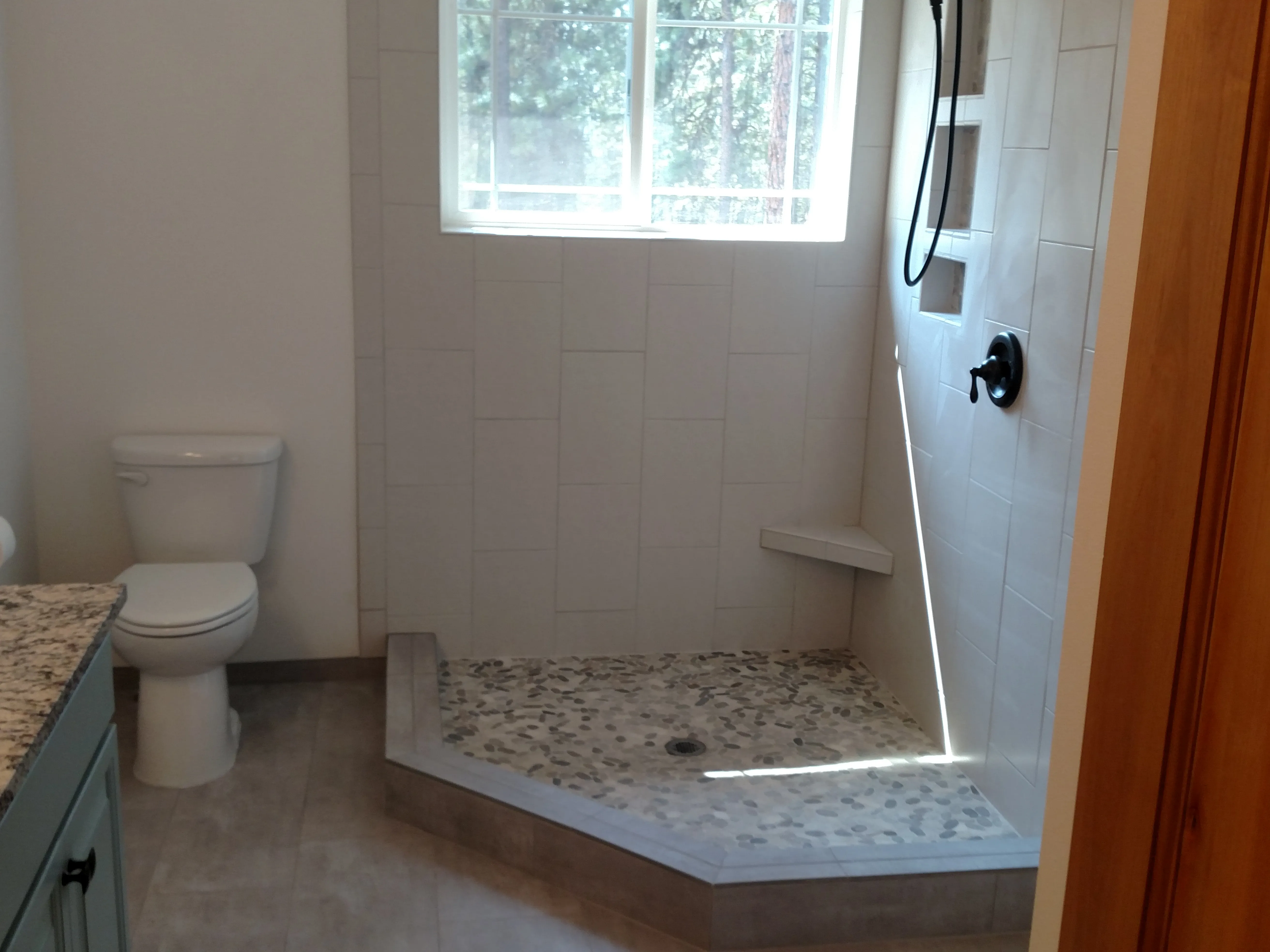 After — Forest-view bathroom remodel. Before: gutted to studs with new blue PEX plumbing, old corner tub left in place. During: cement board (Durock) shower base built up under window. After: finished walk-in shower with white linear tile, pebble-stone shower floor, blue vanity, granite counter, knotty alder door, and the same forest window.